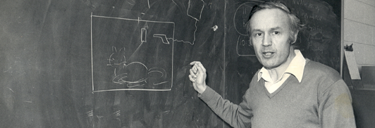 Tony Leggett standing next to his hand-drawn illustration of the Schroedinger thought experiment (notice the cat and gun).