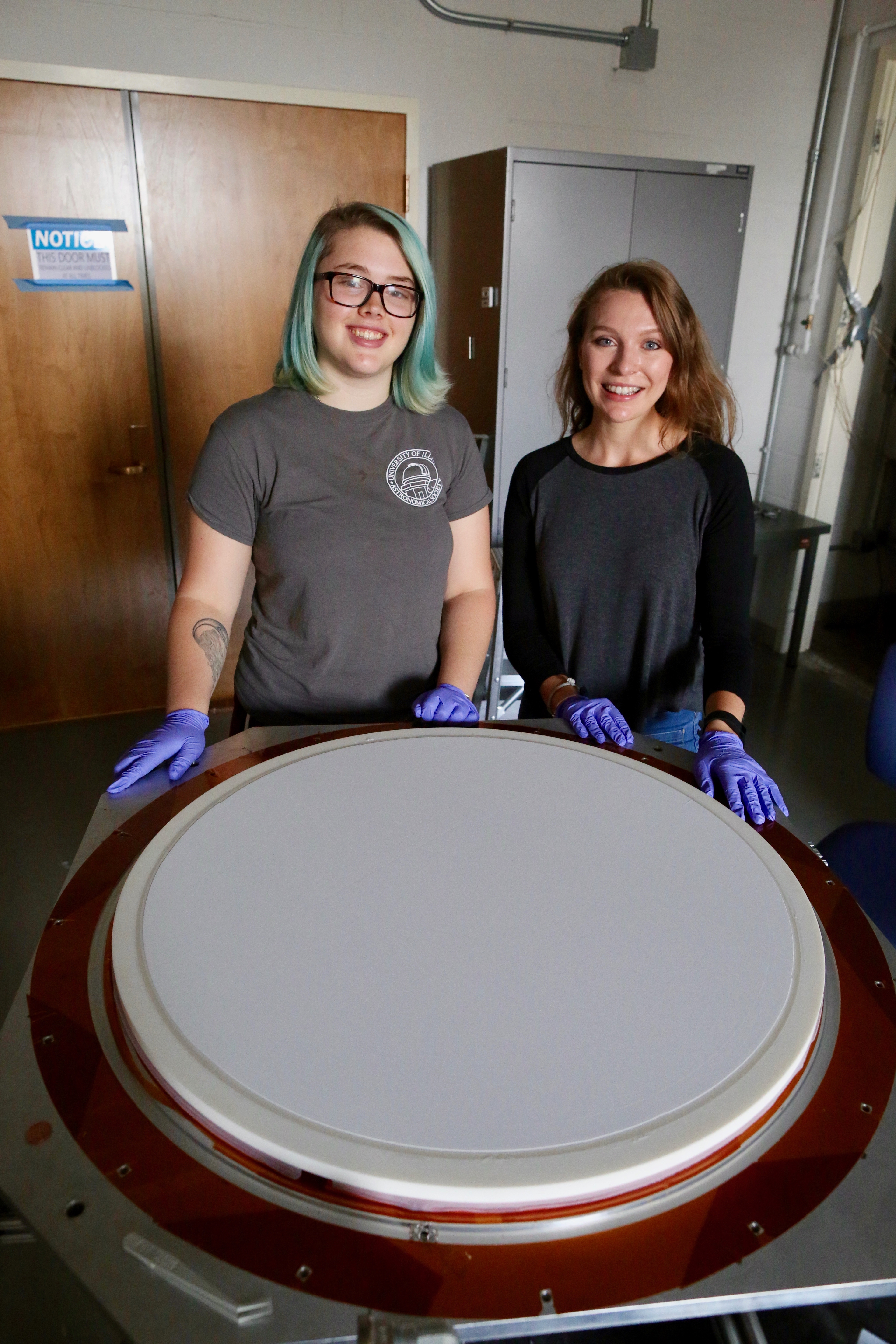 U of I physics undergraduate student Anna Koffman poses with REU student Sierra Barone from Wheaton College, in the Vieira lab, summer 2017. In the foreground is a large lens with a specially developed anti-reflection coating for the South Pole Telescope 3G Camera. Koffman is now a graduate student at the University of Pennsylvania.