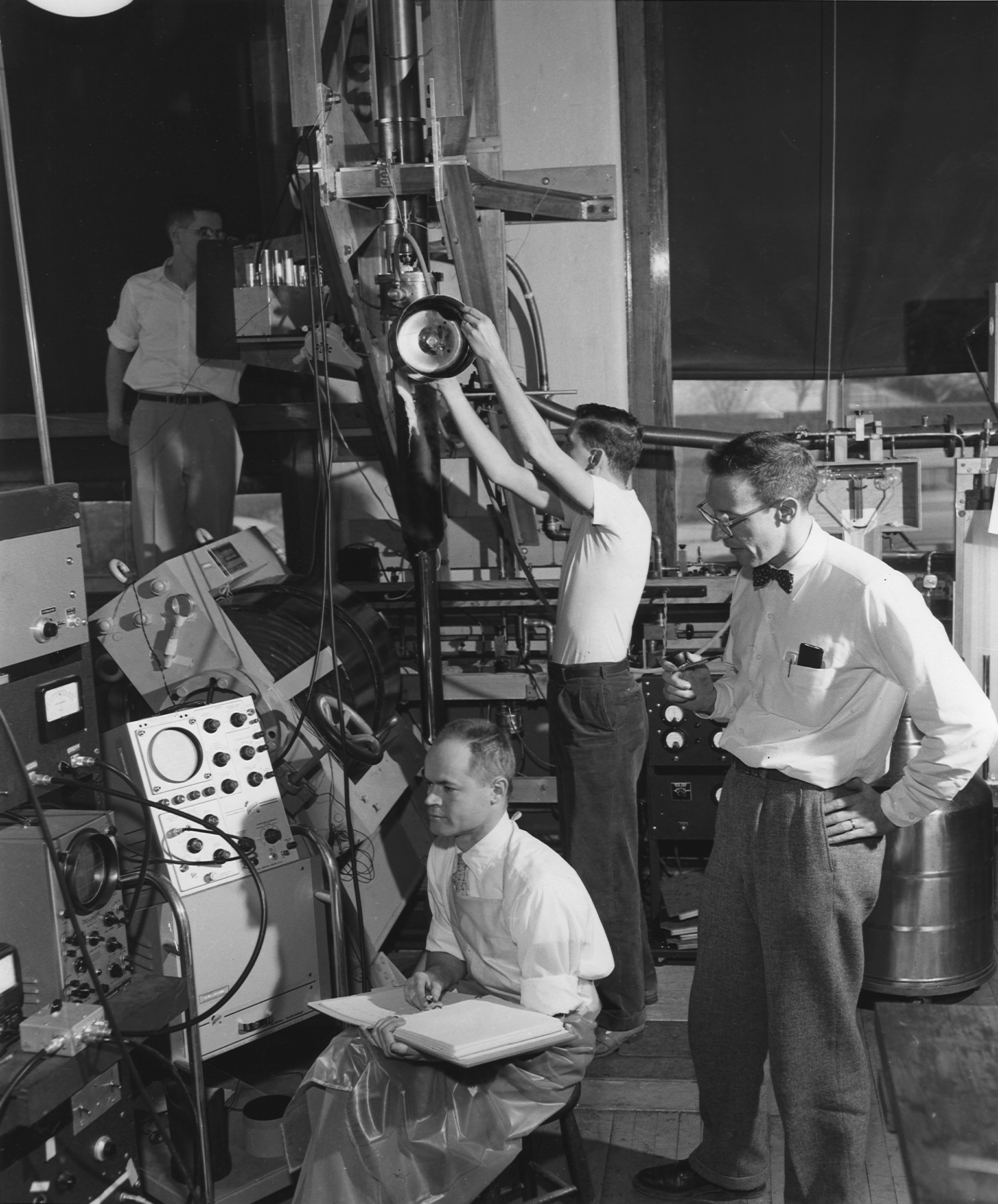 Wheatleyâ€“Mapother low-temp lab, 1957

From the Illinois Alumni News, Vol. 36, No. 3 (April 1957). 'This apparatus on the first floor of the Physics Laboratory looks complex. It doesn't seem so to these physicists: at right, Prof. Dillon E. Mapother; seated with notebook, Prof. J.C. Wheatley; standing in the center of the picture, Thomas Estle, and at back and left, Howard Hart. Estle is an Eastman Kodak fellow and Hart a National Science Foundation fellow. The apparatus includes a cryostat in which temperatures within a few thousandths of one degree of absolute zero (about 459 below zero Fahrenheit) are produced by a process know as adiabatic demagnetization. At low temperatures, materials take on very unusual properties, the study of which often gives special insight into the internal structure, forces and processes in nature.'

Please credit the photo to: University of Illinois Alumni Association Archives
