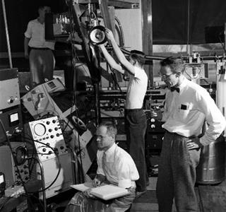 From the Illinois Alumni News, Vol. 36, No. 3 (April 1957). 'This apparatus on the first floor of the Physics Laboratory looks complex. It doesn't seem so to these physicists: at right, Prof. Dillon E. Mapother; seated with notebook, Prof. J.C. Wheatley; standing in the center of the picture, Thomas Estle, and at back and left, Howard Hart. Estle is an Eastman Kodak fellow and Hart a National Science Foundation fellow. The apparatus includes a cryostat in which temperatures within a few thousandths of one degree of absolute zero (about 459 below zero Fahrenheit) are produced by a process know as adiabatic demagnetization. At low temperatures, materials take on very unusual properties, the study of which often gives special insight into the internal structure, forces and processes in nature.' Photo courtesy University of Illinois Alumni Association.