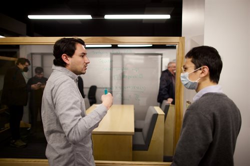 Two graduate students are working at a transparent glass board. Through the glass, tables and chairs are visible, and visitor cubicles are just beyond those.
