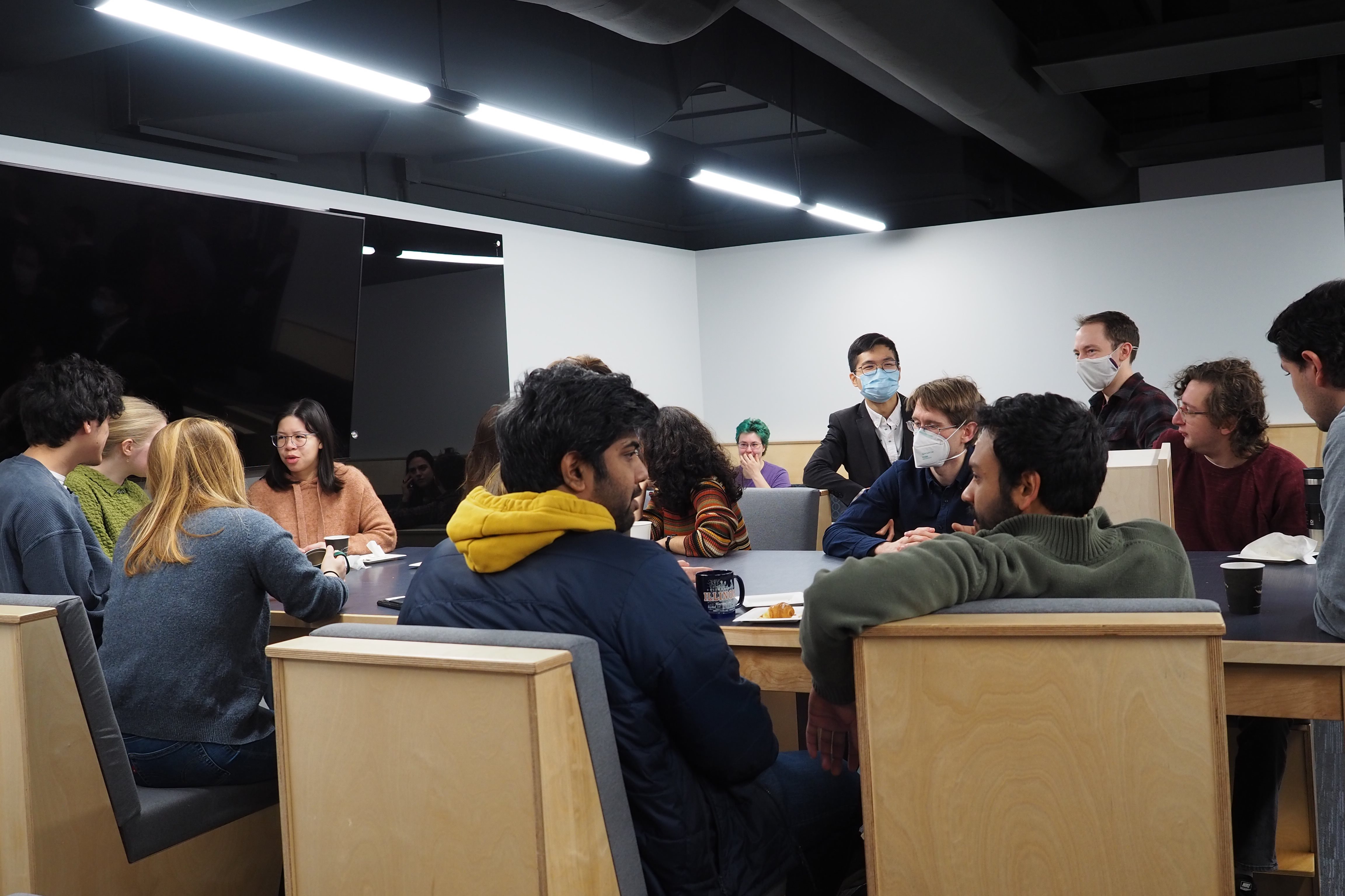 Students and postdocs are gathered around a large table talking and drinking coffee. On the wall is a large monitor and a dark glass dry-erase board.