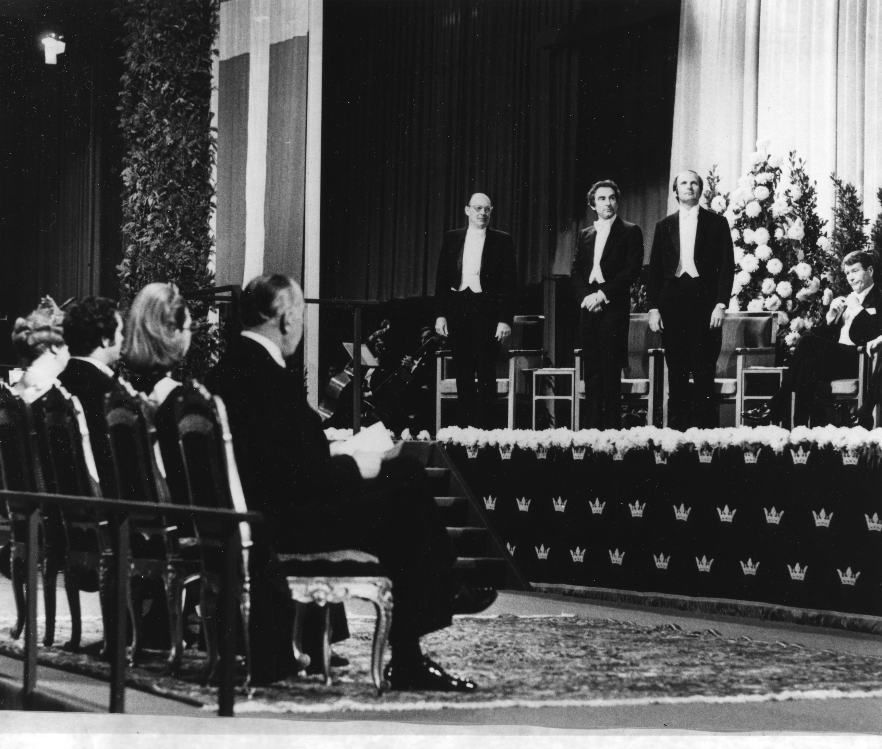 Left to right on stage at the Nobel Prize ceremonies stands John Bardeen, Leon N. Cooper, and John Robert Schrieffer. Others in the photograph are unidentified.