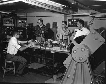 George Newell, Jr., assistant research professor; Theodore Castner, graduate student (PhD, 1958); Louis Hebel, graduate student (PhD, 1957); and Charles Slichter, professor of physics work with equipment in Slichter's basement laboratory in the old Physics Building at the University of Illinois Urbana-Champaign. Their superconducting magnet is shown in the right foreground of the photo.