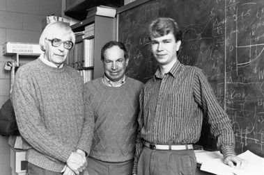 A historical photo of three men standing in front of a blackboard with calculations on it.