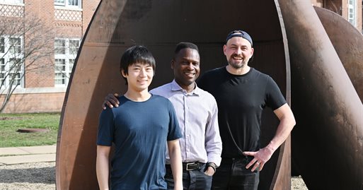 A professor and two graduate students pose in front of a sculpture.